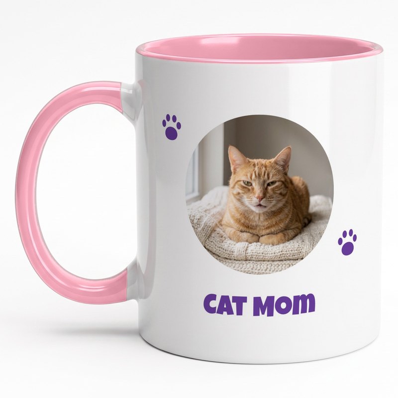 A pink-rim "Cat Mom" mug with an orange tabby cat on a cream blanket in a circle and paw prints